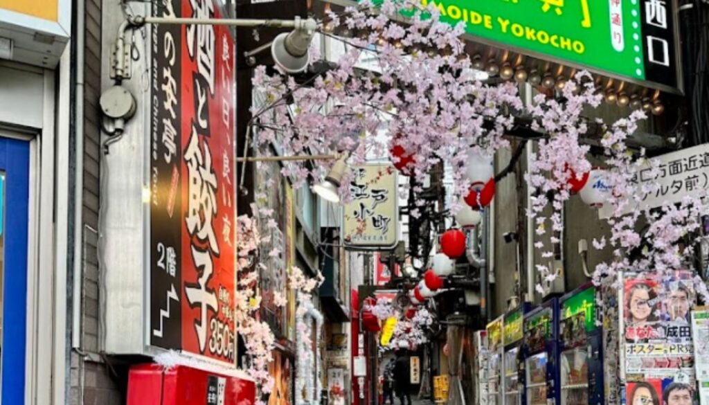 A narrow, vibrant alleyway in Tokyo, Japan, adorned with cherry blossoms, colorful lanterns, and neon signs.