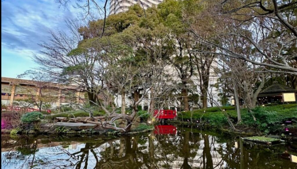 A tranquil Japanese garden with a pond reflecting surrounding trees, a red bridge, and a traditional wooden gazebo. Tall modern buildings rise in the background under a blue sky.