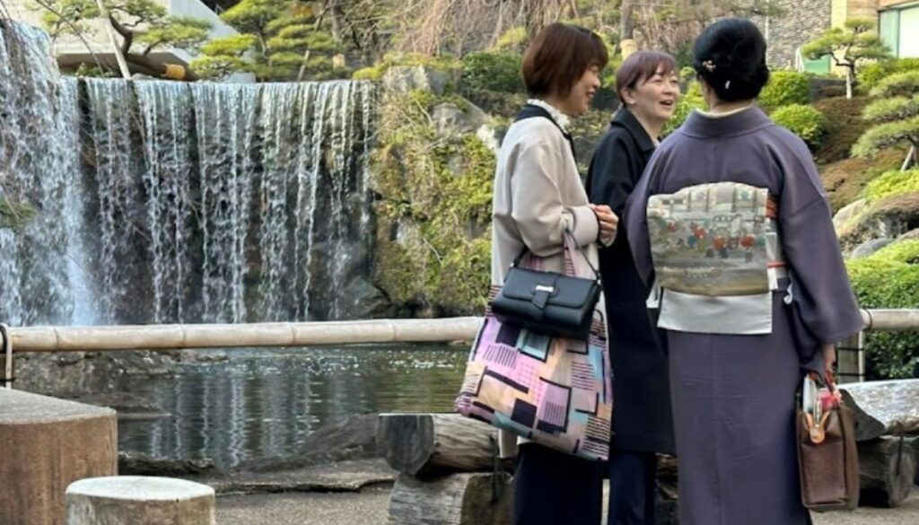 A serene scene of three women standing near a waterfall in a traditional Japanese garden. One woman is wearing a purple kimono with a decorative obi, while the others are dressed in modern clothing. The background features cascading water and lush greenery.