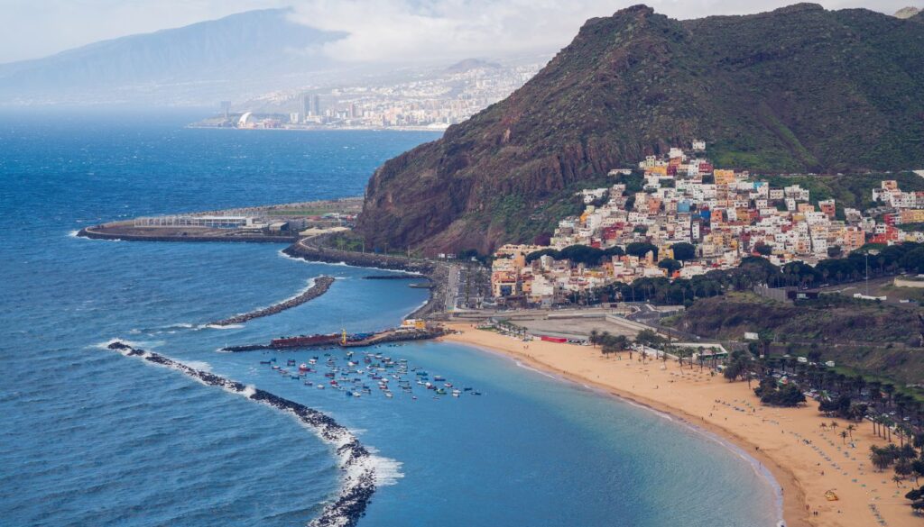 A scenic view of Tenerife's coastline, featuring golden sandy beaches, colorful hillside houses, a vibrant marina, and lush green hills overlooking the ocean.