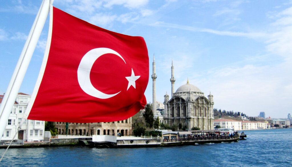 Turkish flag waving near a waterfront with the iconic Ortaköy Mosque in the background, situated by the Bosphorus in Istanbul, Turkey, under a clear blue sky.