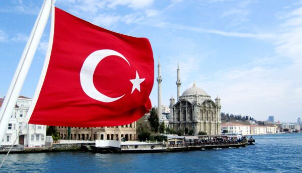 Turkish flag waving near a waterfront with the iconic Ortaköy Mosque in the background, situated by the Bosphorus in Istanbul, Turkey, under a clear blue sky.